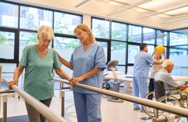 A senior woman assisted by a healthcare professional during a rehabilitation session in a bright, modern facility, while others perform therapy exercises.,Image: 962747604, License: Royalty-free, Restrictions:, Model Release: yes, Credit line: Robert Kneschke/Alamy/Profimedia