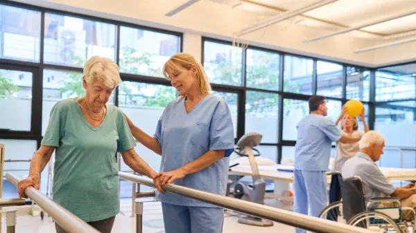 A senior woman assisted by a healthcare professional during a rehabilitation session in a bright, modern facility, while others perform therapy exercises.,Image: 962747604, License: Royalty-free, Restrictions:, Model Release: yes, Credit line: Robert Kneschke/Alamy/Profimedia